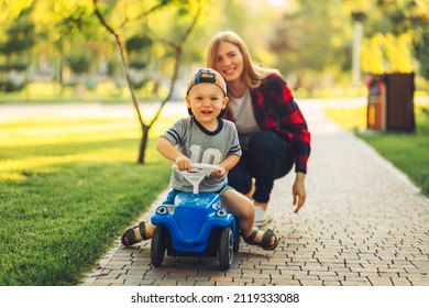 Mom Teaches Her Son To Ride A Toy Car In The Park. Preschooler Driving His Toy Car Outdoors In Summer. Happy Family, Childhood Concept, Outdoor Fun