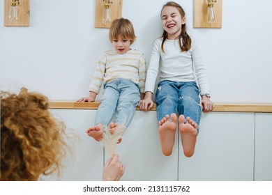 Mom tickling her children's feet with a fluffy cereal spikes. They are sitting on a shelf, a girl is laughing, a boy is slightly afraid.