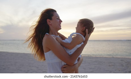 Mom Tosses His Happy Little Daughter In Air On Beach, Having Fun At Sunset. Happy Family And Childhood Concept. Silhouette Of Mother And Healthy Child Flying Over Sun.