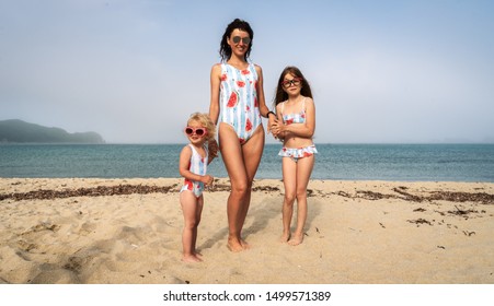 Mom And Two Daughters In The Same Swimsuit On A Sandy Sea Beach. The Younger Daughter With Curly Hair In Pink Sunglasses Looks Away.
