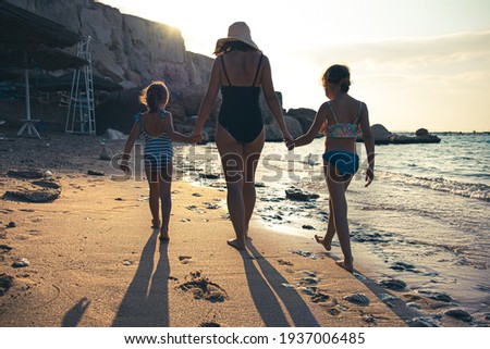 Mom and two little daughters walk along the seashore in swimsuits, holding hands. Vee from the back.