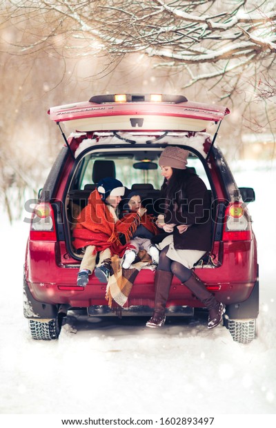 Mom and two sons in a car\
with hot coffee, a winter picnic with rugs and tea, a red car and\
snowfall