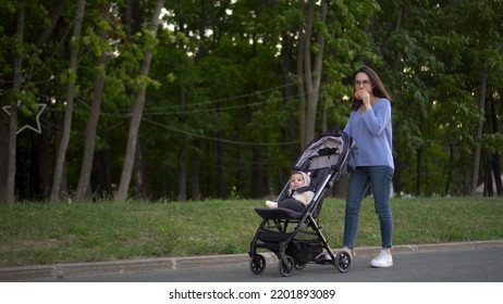 Mom Walks With A Baby In A Stroller And Eats A Burger On The Go. A Young Woman In Glasses Pushes A Stroller With Her Son And Eats A Burger In The Park.