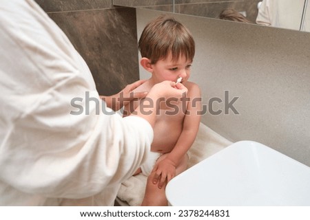 The mom is washing her baby nose with a cotton swab in the bathroom, as part of their daily hygiene routine. Kid aged two years (two-year-old boy)