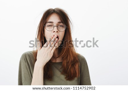 Mom woke up daughter very early. Shot of sleepy attractive caucasian woman with messy brown hair, wearing glasses, feeling tired after night without sleep, yawning, covering opened mouth with palm