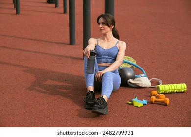 moment to rest, the woman in blue sports gear sits with her water bottle and various exercise tools. maintaining hydration and taking breaks to enhance workout efficiency and safety. - Powered by Shutterstock