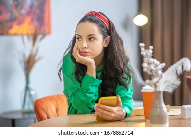 Moment Of Waiting. Beautiful Young Woman With A Yellow Smartphone In Hand, Sitting At A Table In Anticipation, Propping Her Head With Her Hand, Looking To The Side.