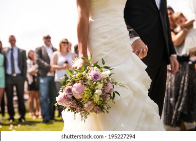 Moment in wedding,  bride and bridegroom holding hands with bouquet and wedding guests in background