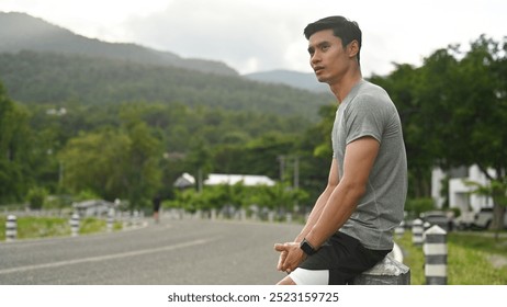 Moments of Reflection: A young man sits on a roadside barrier, taking a moment to rest after a workout, with scenic mountains in the background, reflecting on his fitness journey. - Powered by Shutterstock