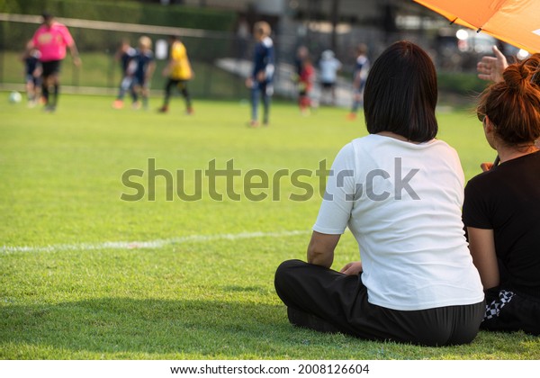 Moms sitting and watching their sons playing\
football in a school tournament on a sideline with a sunny day.\
Sport, outdoor active, lifestyle, happy family and soccer mom and\
soccer dad concepts.