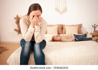 Mom's Tired Of The Baby. A Two-year-old Girl Lying On The Bed Hysterically Screaming Crying. The Woman Is Unhappy, Covers Her Face With Hands And Does Not Want To Listen The Baby Doesn't Want To Sleep