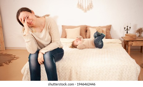 Mom's Tired Of The Baby. A Two-year-old Girl Lying On The Bed Hysterically Screaming Crying. The Woman Is Unhappy, Covers Her Face With Hands And Does Not Want To Listen The Baby Doesn't Want To Sleep