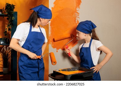 Mon And Child Using Orange Paint On Roller Painbrush To Change Wall Color, Working On House Redecoration. People Having Fun Painting Apartment Dcor Interior, Renovating With Tools And Roll.