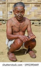 Mon district, Nagaland, India - 11 20 2010 : Outdoor full body front portrait of old Naga Konyak tribe head hunter warrior with traditional facial and chest tattoo sitting on low stool