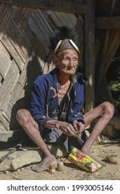 Mon district, Nagaland, India - 11 25 2013 : Outdoor portrait of old Naga Konyak tribe head hunter warrior with traditional facial tattoo wearing hat with boar tusks, sitting and preparing betel quid