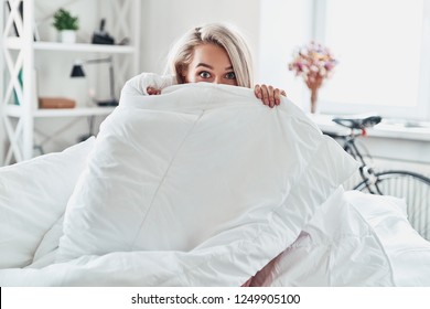 Monday morning. Attractive young woman covering half of her face with blanket and looking at camera while sitting in bed at home