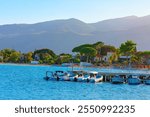 Mondello Beach in Palermo, Sicily Italy. Coastal scene with several boats docked at a pier, set against a backdrop of lush greenery and mountains. Sea water is blue and area appears to be a peaceful