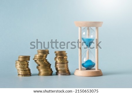 Money accumulation, investment concept - three stacks of golden coins with small sand clock on blue background.