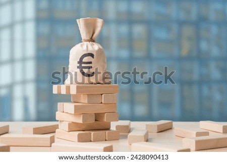 Money bag with Euro symbol on collapsing wooden tower over defocused cityscape. Concept of financial instability, risk, potential downfall of financial investments