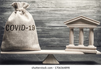 Money Bag With The Word Covid-19 And Government Building On The Scales. State Support For Infected And Sick People. Financial Help. Insurance. Coronavirus Pandemic Infection. Healthcare, Medicine.