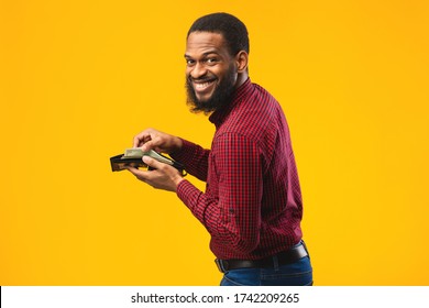 Money Concept. Black Man Holding Open Wallet With Cash And Smiling Over Orange Studio Background, Copyspace