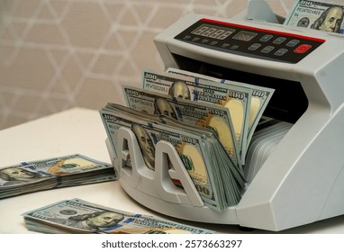 A money counting machine processes a large amount of cash while neatly organized stacks of hundred-dollar bills are placed next to it on a table.