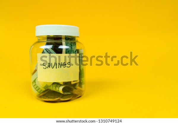 Money jar ful\
of paper bills and coins with savings sign on the sticker. Isolated\
on yellow background. Copy\
space