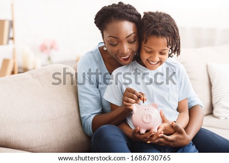 Money Saving Concept. Excited black mom and daughter putting coins into piggy bank, free space