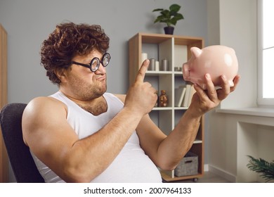 Money savings, economy. Portrait of funny frowning face fat man wagging with raised index finger at piggy bank looking at moneybox showing shaming or arguing gesture feeling lack of cash