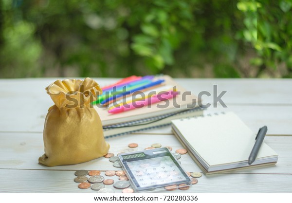 Money savings, mortage and loan\
planning concept. Golden money bag bank, coins, notebook,\
calculator and pens on wooden table and green.garden\
background.