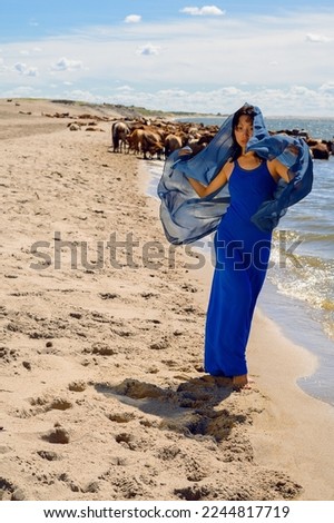 A Mongolian young girl with a blue dress on the beach of the lake is walking behind a herd of horses