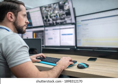 Monitoring. Attentive involved bearded young man with keyboard sitting watching in front of computer screens