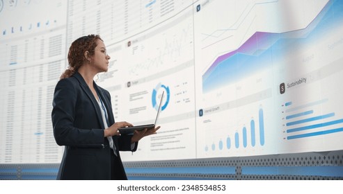 In the Monitoring Office: Successful Hispanic Businesswoman Holding Laptop Computer And Analyzing Data On Big Digital Screen With Graphs and Charts. Female IT Entrepreneur Running Software Company.