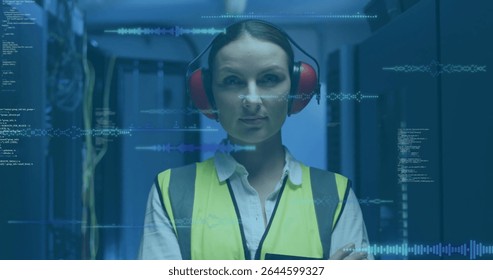 Monitoring tech in high-vis vest red earmuffs scanning racks in blue LED data center, code overlays. Technology, cybersecurity, industrial, engineering, innovation, security, digital