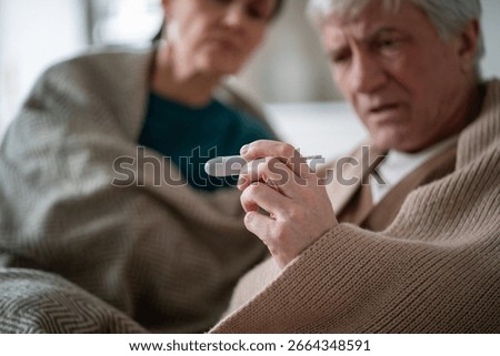 Monitoring temperature during sickness. Senior couple under cover due to illness, shared high temperature. Contagious disease, caregiving in old age, mutual support during illness, prolonged recovery