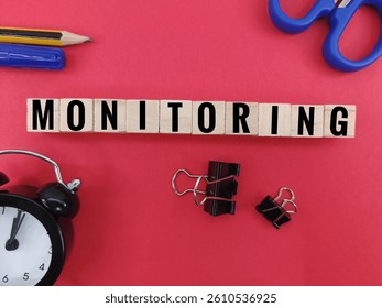 Monitoring word on wooden blocks with office supplies on red background – business management concept, analysis, control, supervision, and productivity planning. 