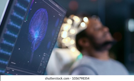 Monitors Show EEG Reading And Graphical Brain Model. In The Background Laboratory Man Wearing Brainwave Scanning Headset Sits In A Chair With Closed Eyes. In The Modern Brain Study Research Laboratory