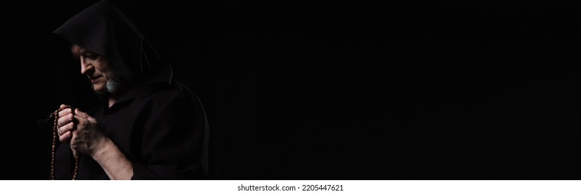 Monk In Dark Hooded Cassock Praying With Rosary Isolated On Black, Banner