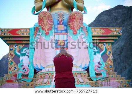 A monk holding hands & praying in front of a giant Maitreya Buddha statue, in North India.
