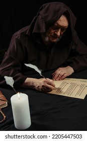 Monk In Hooded Robe Writing On Parchment Near Burning Candle At Night Isolated On Black