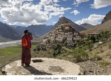 a monk taking picture with mobile phone of ki monastery / key monastery, spiti valley, himachal pradesh, india, surrounded by mountain and river