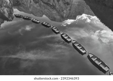 Monochrome, artistic, perpendicular view of wooden boats in row among mirroring peaks and sky on a lake Lago di Braies in the Dolomites. Ideal for black and white poster projects. - Powered by Shutterstock