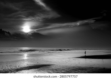 Monochrome beach landscape with dramatic clouds, ocean waves, bright sunset, and a lone human silhouette creating a timeless fine art atmosphere