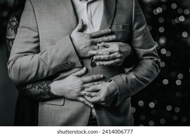 monochrome close-up of a couple's hands clasped around a man in a suit, with the woman's hand wearing a wedding ring.