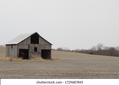 Monochrome Farm Building In Iowa.  Old Shed With Farm Equipment Showing Within Surrounded By A Manicured Field Of Stubble.  Cloudy Overcast Iowa Sky And Solitary Farm Shed With Trees In Background.