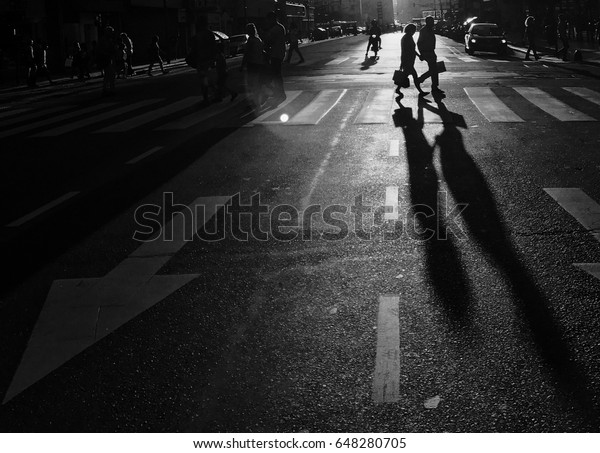 Monochrome\
image of backlit people crossing the\
street