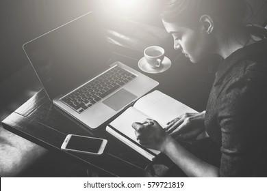 Monochrome Image.Young Business Woman Sitting At Table In Cafe And Writing In Notebook. On Table Is Laptop, Smartphone And Cup Of Coffee. Freelancer Working Outside Office. Student Learning Online.