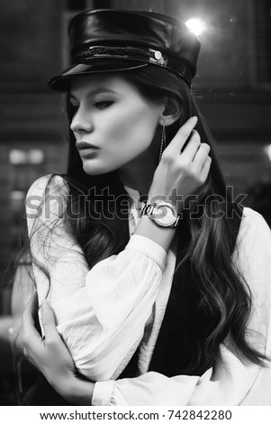 Monochrome outdoor portrait of young beautiful fashionable woman posing in street. Model wearing stylish leather hat, wrist watch, vest, blouse. Lights and reflections on background. Female fashion