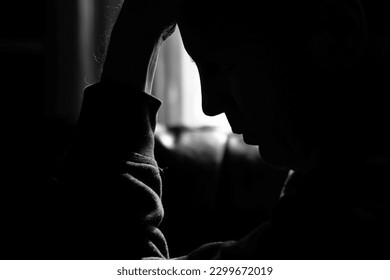 Monochrome photo of a depressed man silhouetted by the light of a window and holding his head in his hands - Shutterstock ID 2299672019