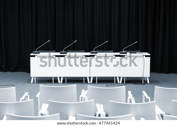 Monochrome picture of empty press conference\
room with seats, stand table and\
microphones